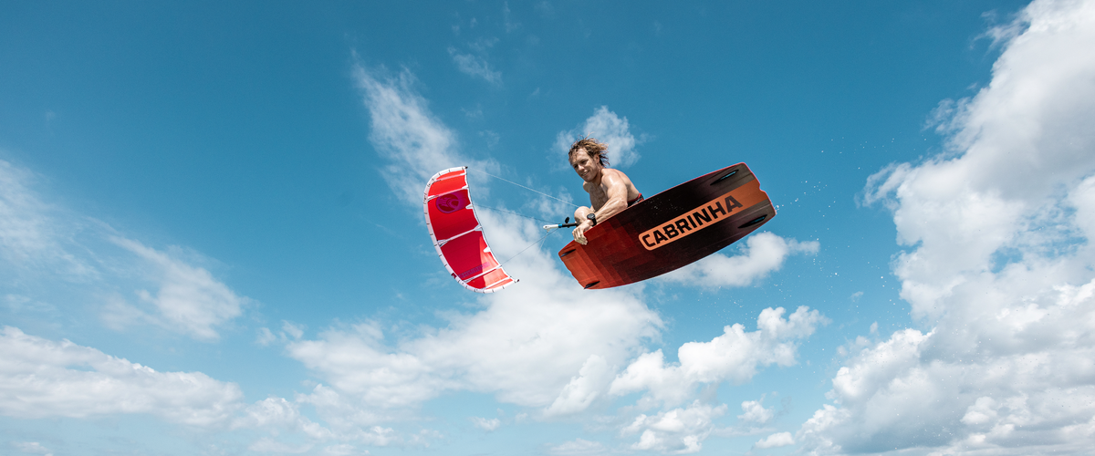 Cabrinha team rider Alberto Rondina performing an aerial board grab while kitesurfing with a red Cabrinha Switchblade against a blue sky.