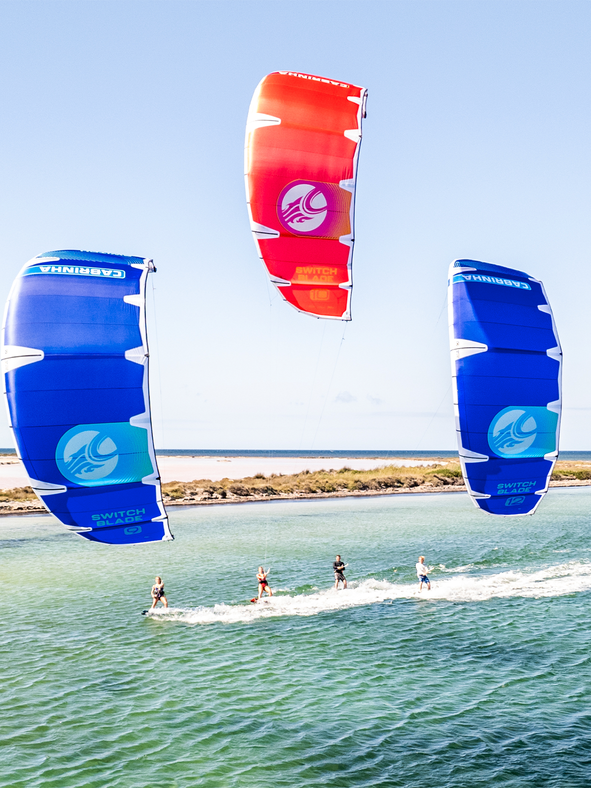 Three Cabrinha Switchblade kites flying above kitesurfers riding across turquoise water on a sunny day.
